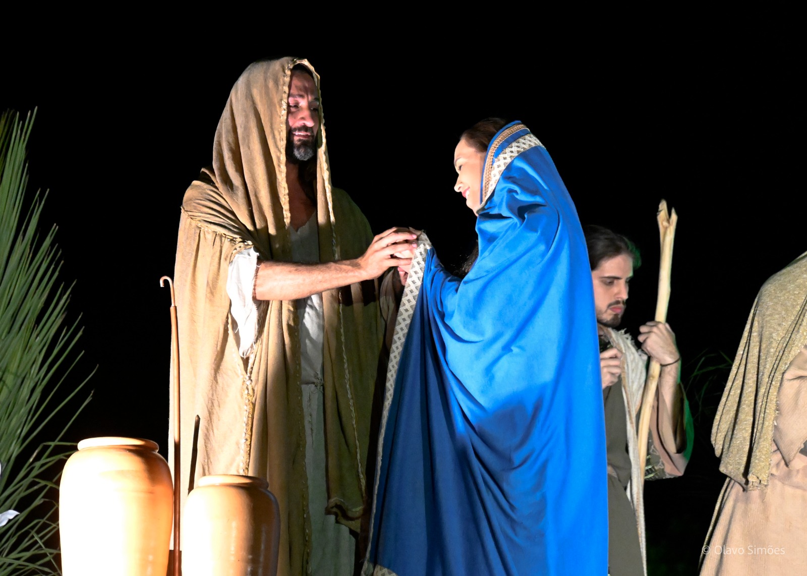 Um ator interpreta Jesus no espetáculo A Paixão de Cristo. Ele está vestido com um manto cor de areia que cobre os seus cabelos e contracena com uma atriz, que também tem os cabelos cobertos por um manto na cor azul. Os dois estão frente a frente e na cena Jesus aparece sorridente, segurando as mãos da mulher, que também está sorridente.