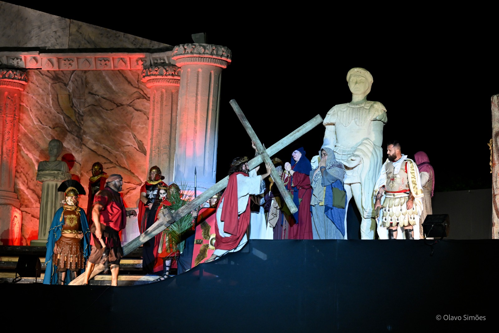 Um grupo de atores interpreta, em um palco, a cena do espetáculo Paixão de Cristo. Ao centro, um homem que interpreta o personagem Jesus carrega uma cruz apoiada nos ombros. Ele é cercado homens caracterizados como soldados romanos e duas mulheres que choram diante da cena. O cenário, ao fundo, é composto por uma estátua e uma edificação com temas romanos.