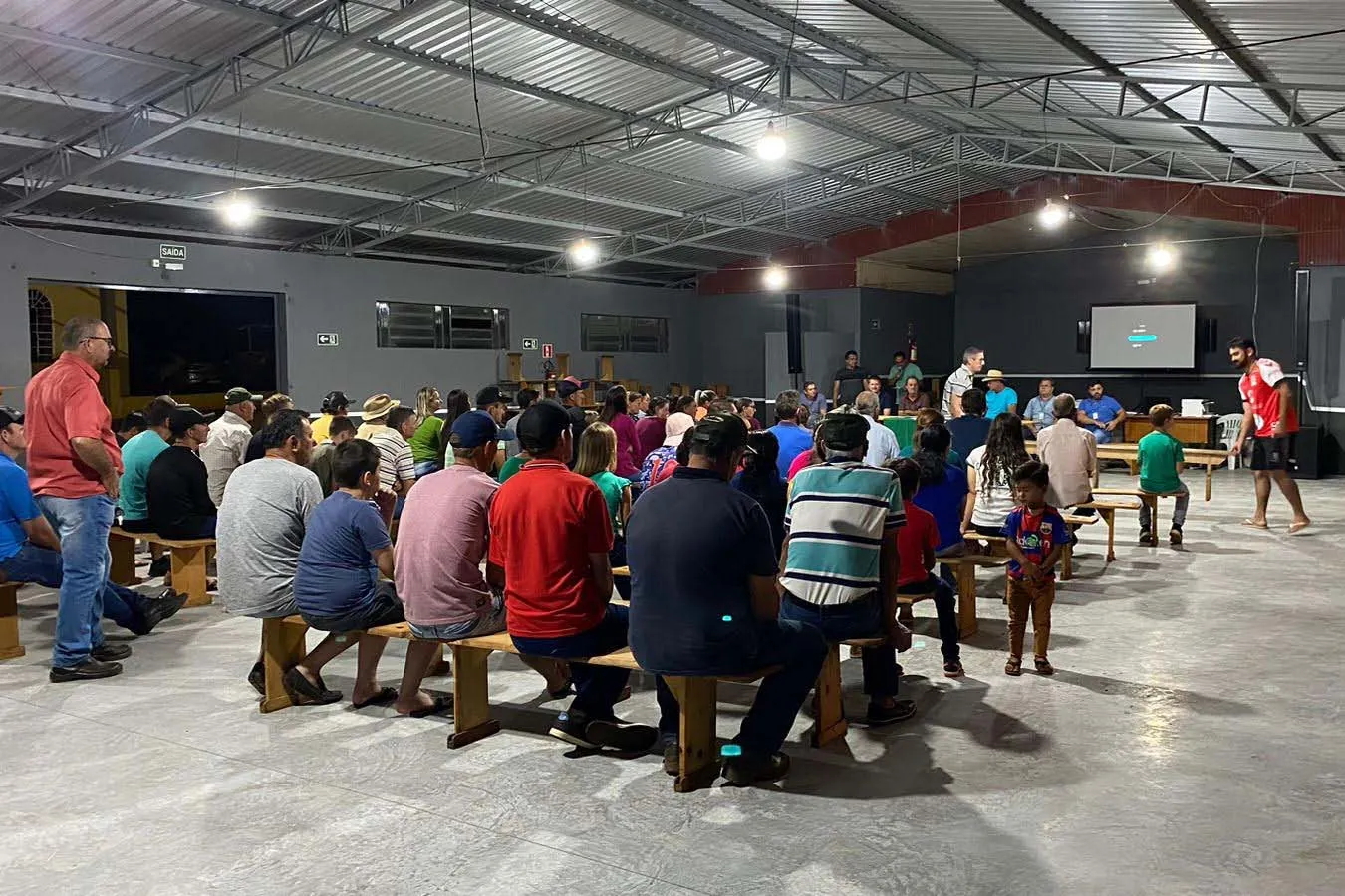Comunidade de pessoas reunidas em um grande galpão, ouvindo uma palestra.