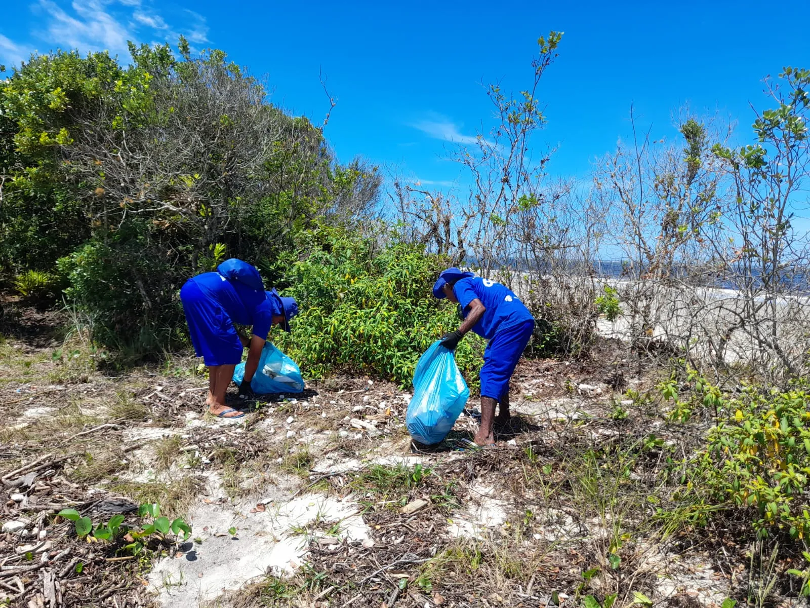 Dois homens limpando a areia da praia com o mar ao fundo