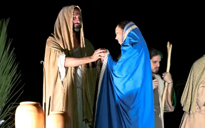 Um ator interpreta Jesus no espetáculo A Paixão de Cristo. Ele está vestido com um manto cor de areia que cobre os seus cabelos e contracena com uma atriz, que também tem os cabelos cobertos por um manto na cor azul. Os dois estão frente a frente e na cena Jesus aparece sorridente, segurando as mãos da mulher, que também está sorridente.