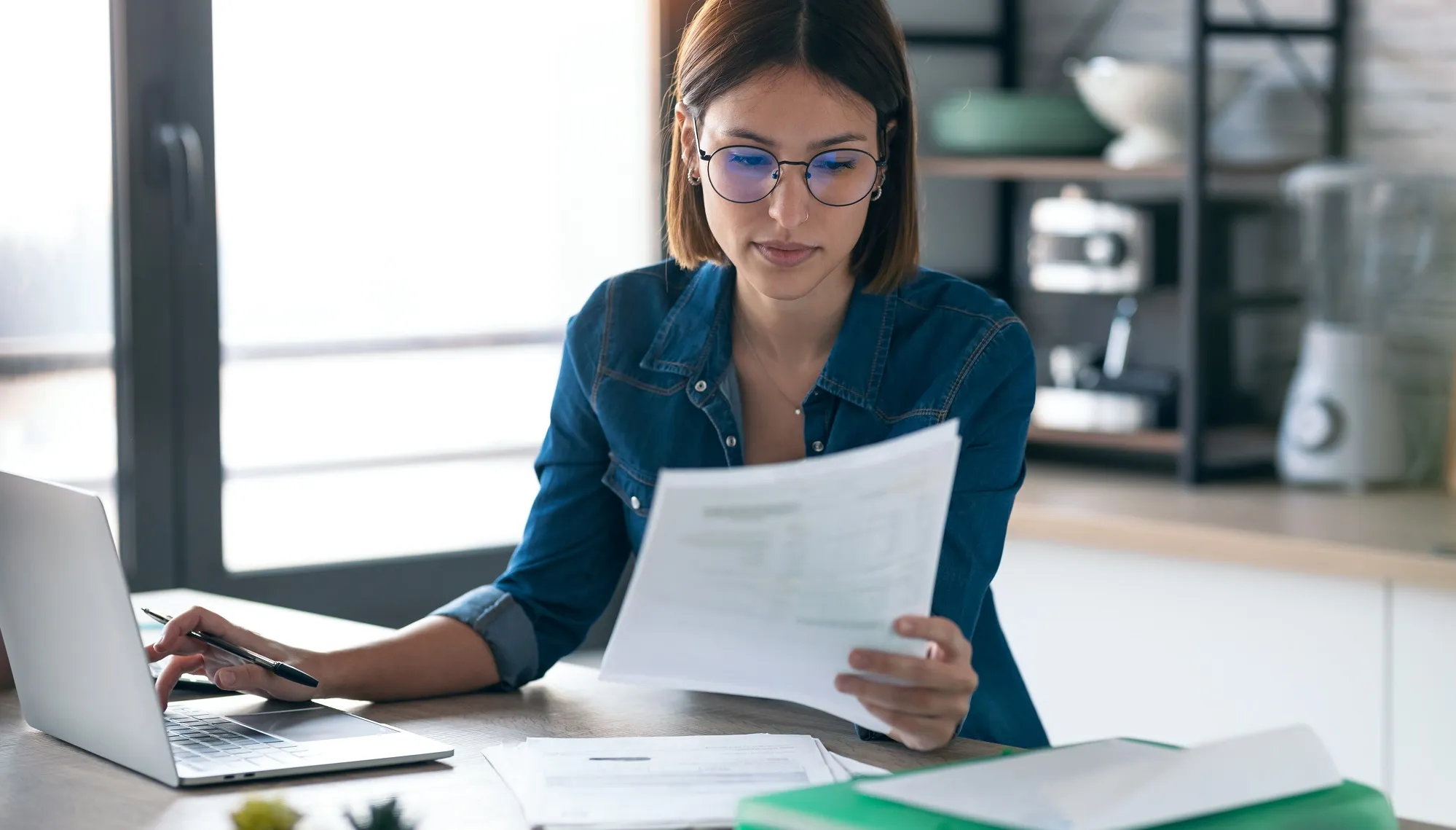 Mulher jovem usando óculos, sentada à mesa da cozinha com o notebook aberto sobre a mesa e analisando uma pilha de documentos de papel