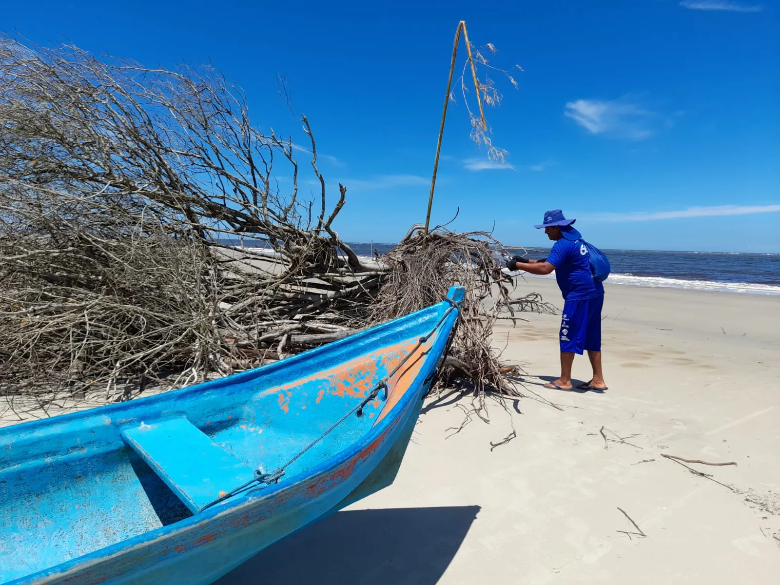 Homem com uniforme da Sanepar limpando a areia da praia do lado de um barco azul pequeno com o mar ao fundo